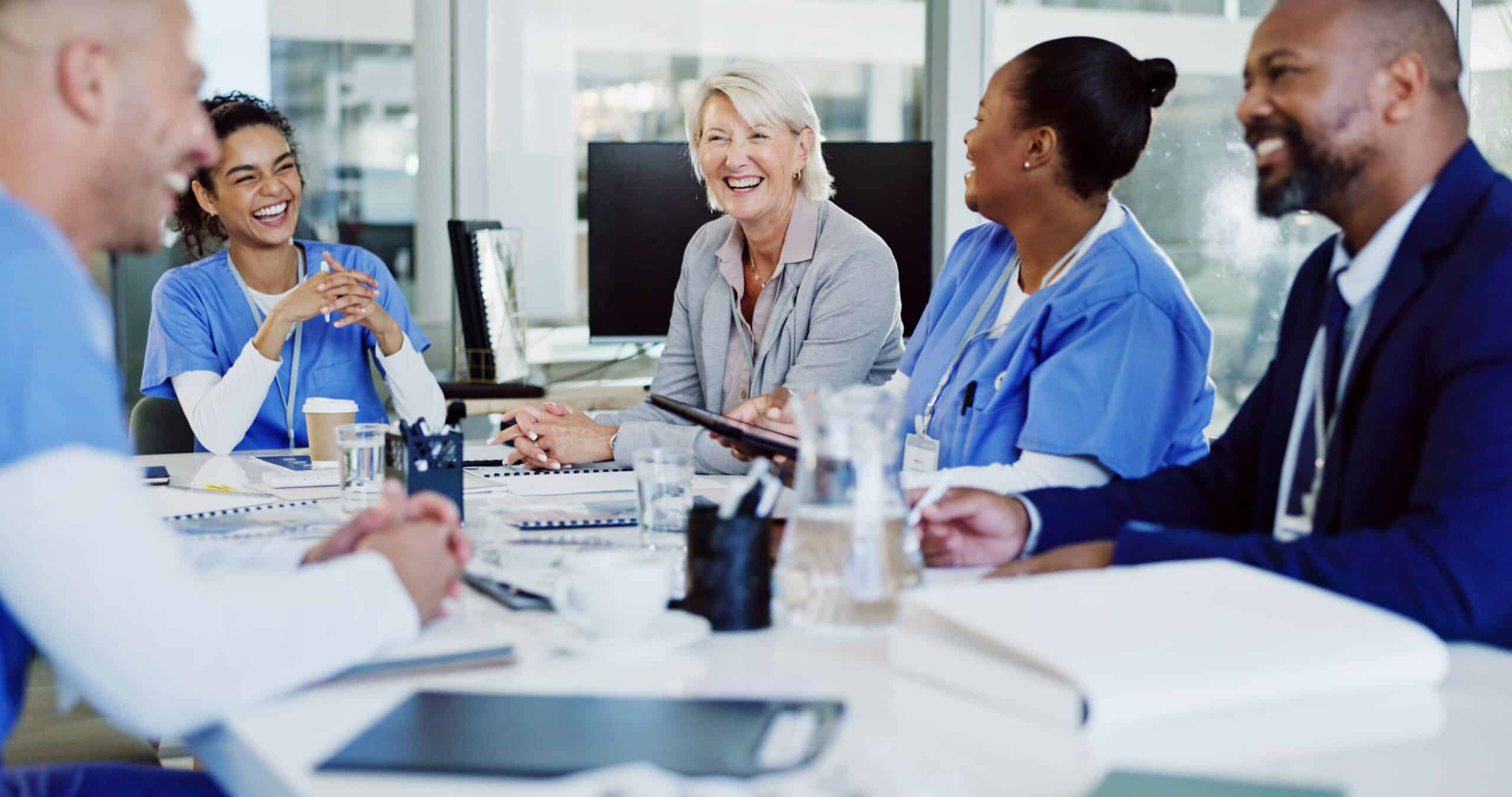 Five professionals, including three in scrubs, discuss urgent care marketing strategy around a meeting table in a bright, cheerful office.