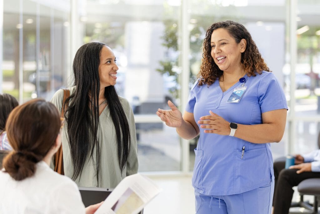 A nurse in blue scrubs discusses urgent care management with a woman in a bright hospital waiting area as another person reads a paper.