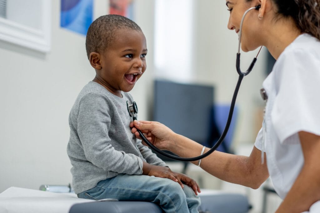 A healthcare professional uses a stethoscope to check a smiling child’s chest during an urgent care clinic marketing photo shoot.