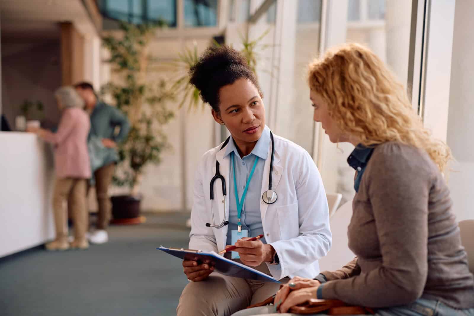 A doctor with a stethoscope talks to a patient while holding a clipboard in a brightly lit waiting area.