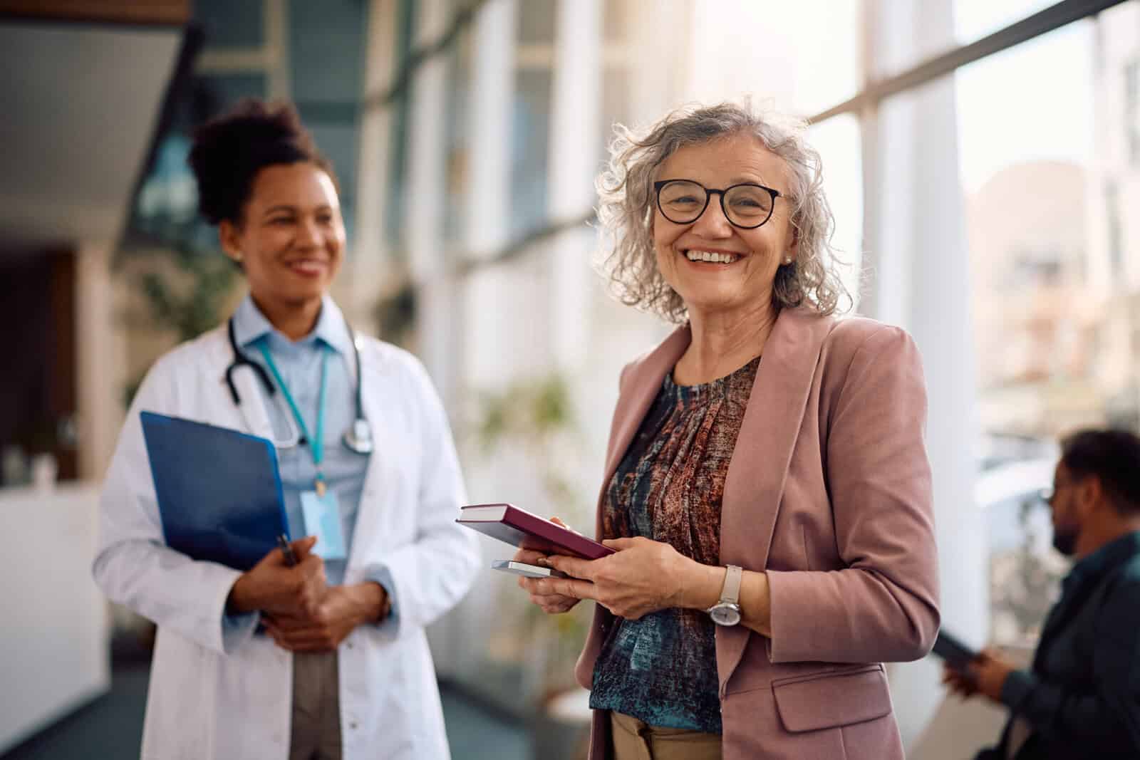 An older woman smiling and holding a notebook stands next to a doctor in a bright, modern office.