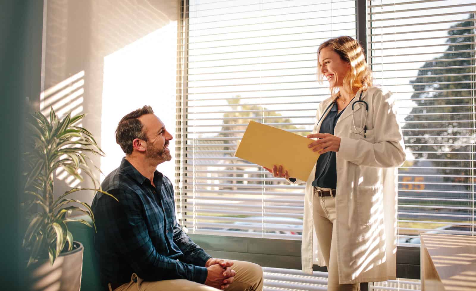 A doctor holding a file stands and talks to a seated patient in a sunlit office with large windows and blinds.